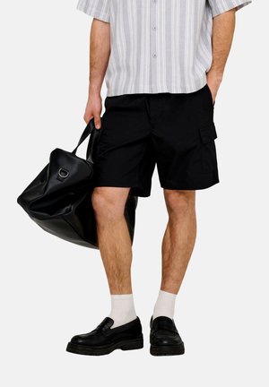 Man wearing black cargo shorts, white socks, black shoes, striped shirt, holding large black leather bag with one hand.
