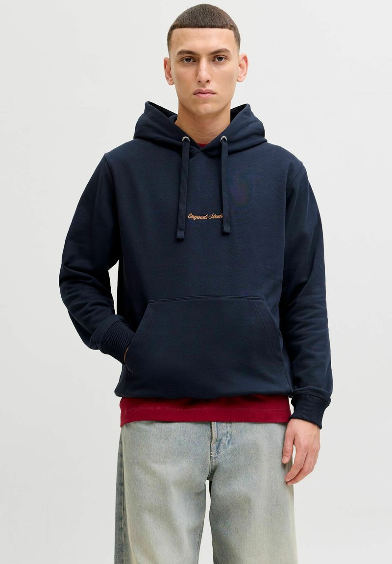 Merineitsi kapuutsiga sviiter puuvilla segu materjalist, ees taskuga ja kuldse tikandiga tekstiga. Kantud punase särgi peal, koos heledate siniste teksadega.