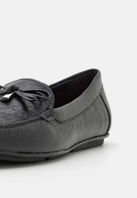 Mocassins noirs en matériau synthétique texturé, avec un nœud sur le devant et une semelle plate et basse pour un style décontracté.