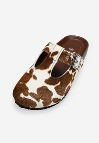 Zuecos con estampado de vaca que presentan un suave y texturizado upper en marrón y blanco, dos recortes frontales y una hebilla plateada sobre una suela oscura y elegante.