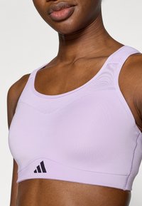 Αθλητικό σουτιέν ανοιχτό μωβ χρώματος από λείο υλικό, με στρογγυλεμένο ντεκολτέ, φαρδιές τιράντες και μαύρο λογότυπο κοντά στη ραφή.