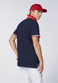 Navyblaues Polohemd mit roten und weißen gestreiften Ärmeln und Kragen, mit einem Logo auf dem Kragen. Modell trägt eine rote Mütze und weiße Hose.