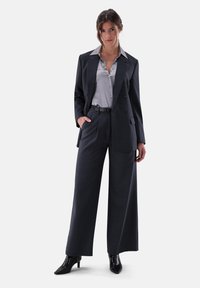 Dunkelgrauer zweireihiger Blazer mit weit geschnittenen Hosen und einem hellgrauen Hemd mit Knöpfen. Glänzende schwarze spitze Absatzschuhe.