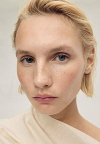 Close-up of a face with light skin, freckles, blue eyes, and short blonde hair. Wearing a draped, textured beige garment. Neutral background.