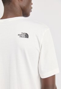 Bijela majica s kratkim rukavima koja ima crni logo "The North Face" na gornjem dijelu leđa. Mekana tkanina s klasičnim krojem i glatkom teksturom.