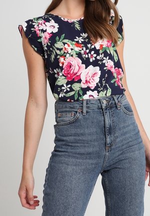 Vrouw draagt een marineblauwe blouse met roze en witte bloemen, gestoken in een hooggetailleerde blauwe spijkerbroek, tegen een effen achtergrond.