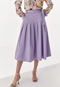 Kvinna i en högmidjad lavendelfärgad midiklänning med veck och en sidobåge, kombinerad med beige högklackade sandaler med remmar runt anklarna och en blommig blus.