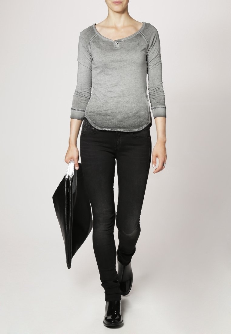 Femme marchant portant un haut gris à manches longues, un jean skinny noir, des bottes noires, portant un grand dossier ou un portfolio noir.