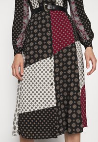 Žena ve vyšívaných midi šatech s páskem, které mají vzorované černé, bílé a burgundské látky a dlouhé rukávy.