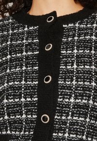 Mustavalkoinen ruudullinen tweed-neule, jossa on pyöreä pääntie ja viisi koristeellista nappia mustilla keskiosilla ja hopeisilla yksityiskohdilla.