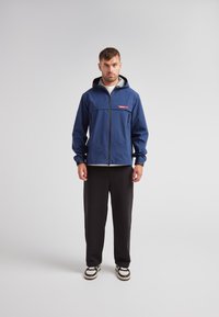 Veste zippée bleue avec capuche, arborant un logo rouge, des poches avant et une texture lisse. Associée à un pantalon noir et des chaussures.