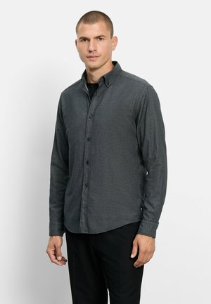 Mann mit kurzen Haaren, der ein dunkelgraues Hemd mit Knöpfen und schwarze Hose trägt, steht vor einem schlichten hellen Hintergrund.
