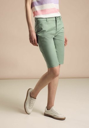 Un short bermuda vert clair avec une coupe ajustée et des poches latérales. Porté avec des baskets blanches, sur un fond neutre.