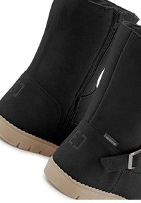 Schwarze Wildleder-Winterstiefel mit einem seitlichen Reißverschluss, flacher brauner Laufsohle, verstellbarer Riemen-Detail und einer strukturierten Oberfläche. Markenetikett auf einer Seite.