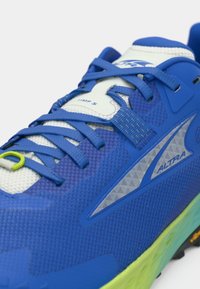 Chaussure de sport bleue avec un dessus en maille texturée, des accents blancs contrastants, des détails réfléchissants et une boucle de lacet verte sur la languette.