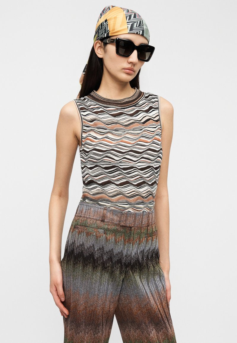 Knit sleeveless top with wavy stripes in black, white, and earthy tones; paired with colorful zigzag-patterned pants with a metallic finish.