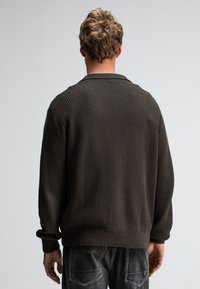 Dunkelgrüner, gerippter Strickpullover mit lockerer Passform, ausgestattet mit einem Kragen, langen Ärmeln und einem gerippten Saum. Getragen mit dunklen Jeans.