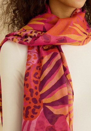 Žena v bielom tričku s výrazným šálom s abstraktnými vzormi inšpirovanými zvieratami vo farbách červenej, oranžovej, ružovej a fialovej, ktoré má prehodené okolo krku a ramien.