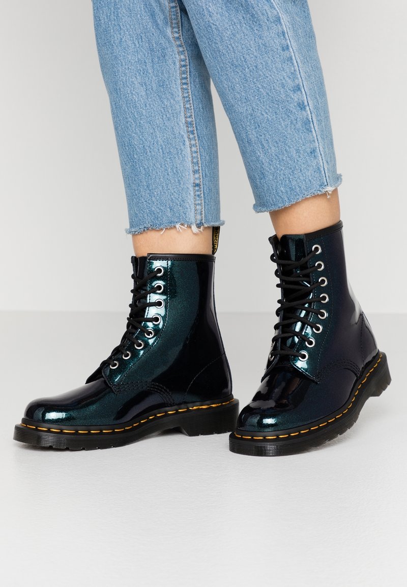 Bottines en cuir verni noir et turquoise avec un design à lacets, des coutures jaunes et une finition brillante. Portées avec un jean bleu court.