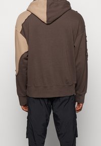 Persona che indossa un hoodie bicolore marrone con il cappuccio alzato, abbinato a pantaloni cargo neri larghi, mostrata di spalle su uno sfondo neutro.