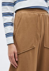 Bruine joggingbroek met een zachte textuur en een elastische tailleband, voorzien van grote zijzakken en een hand van de drager die op de stof rust.