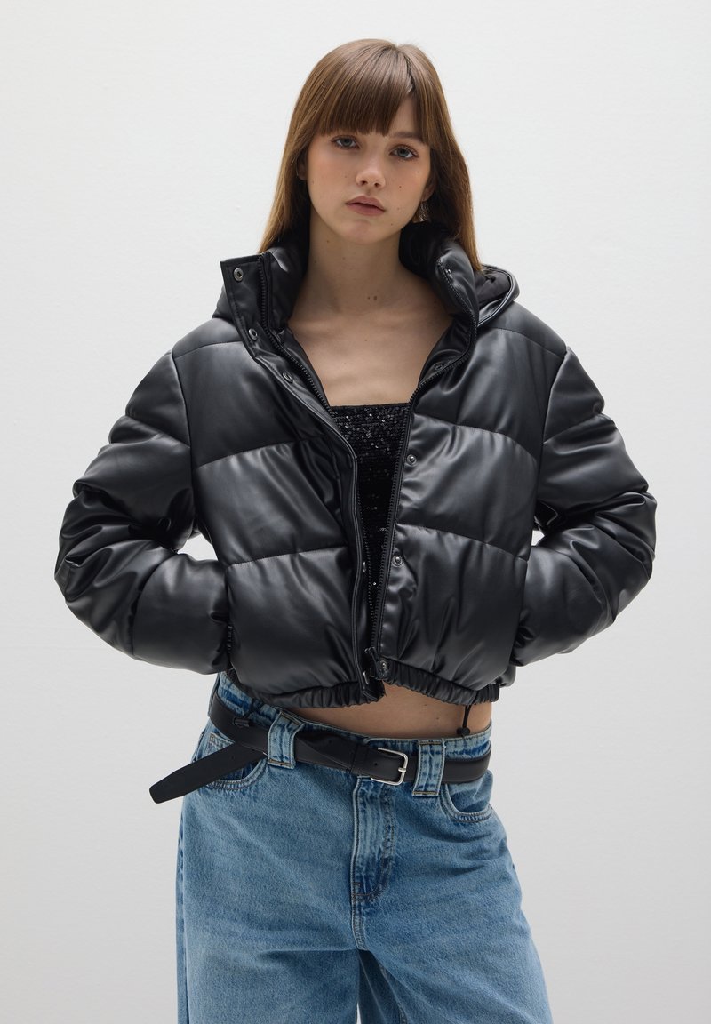 Veste en tissu synthétique noir, style doudoune, avec un design court, fermeture éclair, col montant et poignets élastiqués. Associée à un jean taille haute.