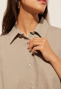 Camisa de lino beige con cuello y botones perlados, que muestra una textura suave. Mano visible ajusta un botón cerca del área del cuello.