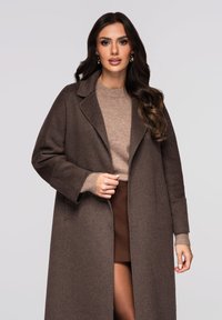 Brun uldfrakke med hakkede revers, overdimensioneret silhuet, kombineret med en lysebrun sweater med en diskret tekstur og neutrale toner.