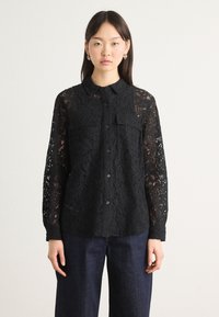Black lace shirt with long sleeves, button-down front, two chest pockets, and intricate floral pattern, paired with dark denim trousers.