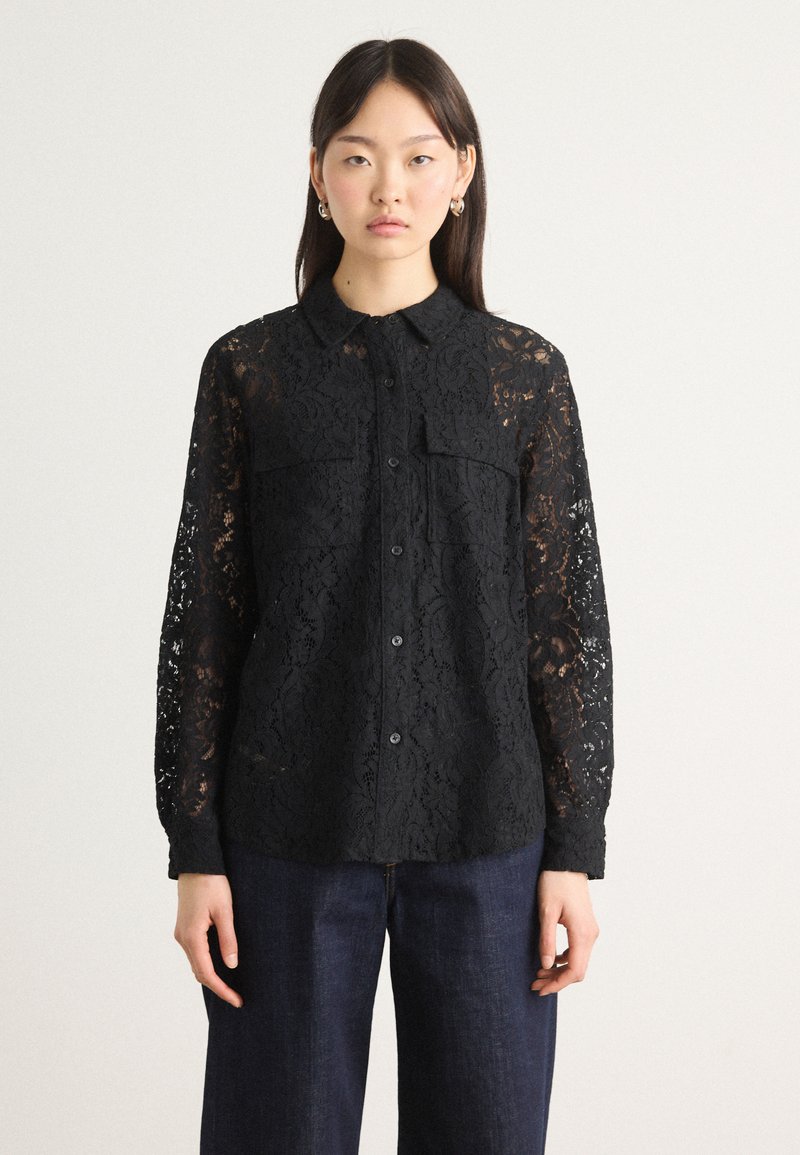 Black lace shirt with long sleeves, button-down front, two chest pockets, and intricate floral pattern, paired with dark denim trousers.