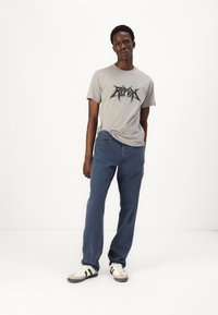 Grå t-shirt med sort grafisk design, parret med blå jeans. Modellen bærer hvide sneakers med sorte striber. Enkel, afslappet outfit.