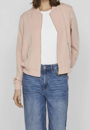 Persoon draagt een lichtroze jas met ritssluiting over een witte blouse en blauwe spijkerbroek, staand tegen een effen witte achtergrond.