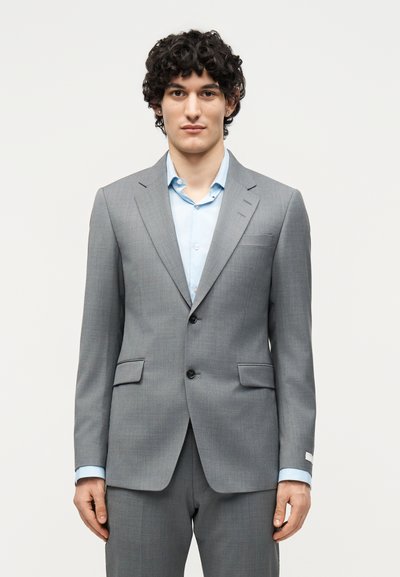 Άνδρας με σγουρά σκούρα μαλλιά, φορώντας ανοιχτό μπλε πουκάμισο και προσαρμοσμένο γκρι σακάκι με δύο κουμπιά, στέκεται μπροστά σε λευκό φόντο.