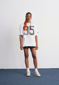 T-shirt oversize blanche avec un graphique noir "95", associée à un short noir et des baskets claires. L'arrière-plan est uni et clair.