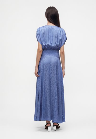 Femme aux longs cheveux foncés portant une robe maxi fluide bleue à motifs et des sandales noires à talons, debout, dos tourné sur un fond blanc.