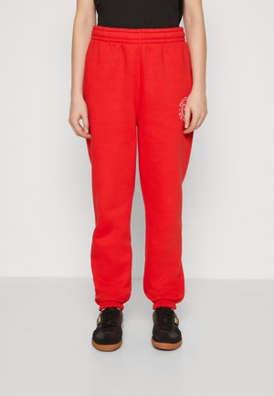 Personne portant un pantalon de survêtement rouge vif avec une taille et des chevilles élastiques, associé à des baskets noires et un haut noir, debout devant un fond uni.