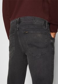 Jean Lee gris foncé porté par une personne avec la main dans la poche, haut bordeaux visible, poches arrière et patch logo Lee à la taille.