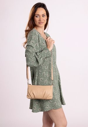 Helle beige Leder-Crossbody-Tasche mit gerafftem Design, verstellbarem Riemen und subtilen metallischen Akzenten, kombiniert mit einem gemusterten grünen Kleid.