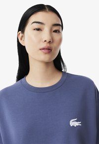 Sweatshirt di cotone viola con colletto a giro e un piccolo logo di coccodrillo bianco sul lato sinistro del petto. Texture liscia.