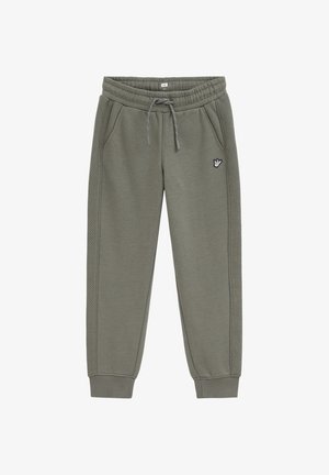 Pantalon de jogging vert olive avec taille à cordon, poches latérales, poignets côtelés, motif matelassé sur les côtés et petit patch avec un geste de la main sur la cuisse.
