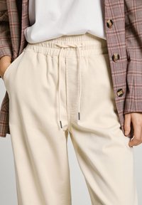 Pantalones de color crema, con cintura elástica y cordón. Textura lisa, diseño de pierna ancha y bolsillos laterales. Combinados con una chaqueta de cuadros.