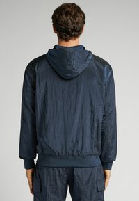 Marinblå lätt jacka med huva, med en krinklad textur och ribbade ärmslut och nederkant, designad för en bekväm passform.