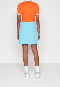 Osoba stojící zády, oblečená v oranžovo-modrém límečkovém tričku, světle modré sukni, bílých ponožkách a bílých teniskách.