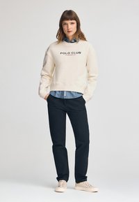 Felpa crema con il testo "POLO CLUB", indossata sopra una camicia di jeans azzurro chiaro, abbinata a pantaloni scuri e scarpe da ginnastica chiare.