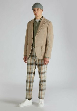Beige corduroy blazer, olijfgroen shirt eronder, geruite broek in roomwit, bruin en blauw, gecombineerd met witte sneakers en een grijze gebreide beanie.