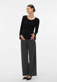 Femme enceinte en haut à manches longues noir avec un nœud et un large pantalon gris, debout, souriante et tenant son ventre.