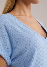 Haut en tricot bleu avec un col en V et des manches courtes, doté d'un motif en chevrons texturé et d'un matériau léger et respirant.