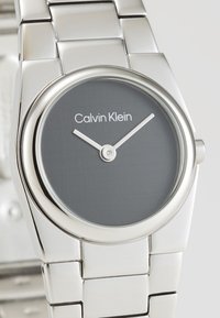 Silverfärgad rostfri ståluhr med en rund grå urtavla, minimalistisk design och Calvin Klein-märke. Utrustad med stiliga tim- och minutvisare.