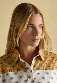 Blond naine, kellel on sinised silmad ja kes vaatab paremale, kannab lillemustrilist pluusi, millel on kollased ja valged osad. Neil on neutraalne beež taust.