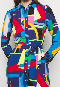 Flerfärgad skjortklänning med geometriska mönster i blått, rött, gult, grönt och vitt. Tyget är slätt med midjebälte.